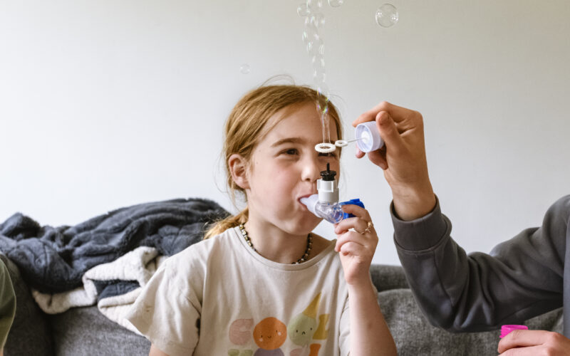 Child blows through a medical device, with her parent holding a bubble maker in front of the output, which makes bubbles.