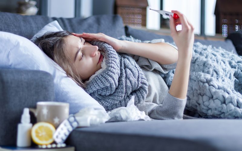 Sick exhausted girl in scarf is lying in bed wrapped in blanket. Young woman with fever and headache is measuring temperature with thermometer, treated at home. Winter cold and flu concept.